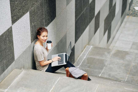 Positive young Vietnamese woman sitting on steps with cup of coffee and laptop when doing homework for college classの写真素材