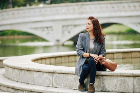 Elegant smiling young woman sitting at fountain in city park and looking at riverの写真素材