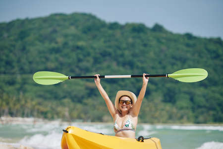 Smiling excited young woman sitting in kayak boat and rising hands with paddleの写真素材