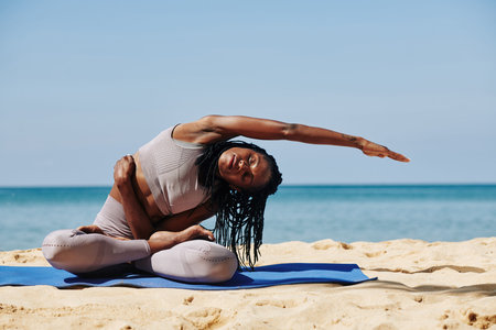 Young sporty woman sitting on yoga mat in lotus position and leaning to the left to stretch her...
