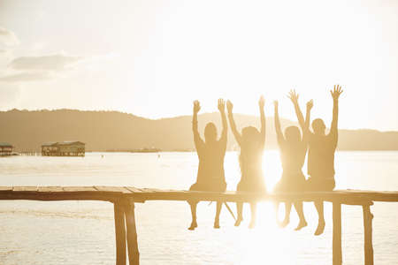 Group of happy excited friends sitting on handmade wooden pier, shouting and raising arms in sunset sun rays, backlitの写真素材