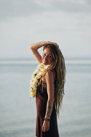 Portrait of smiling beautiful woman with dreadlocks posing with flower necklace against calm seaの写真素材