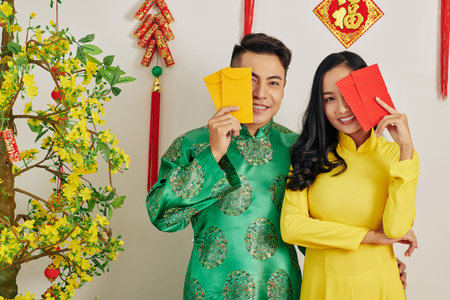 Hugging young Asian couple in traditional Tet costumes standing with lucky money envelopesの写真素材