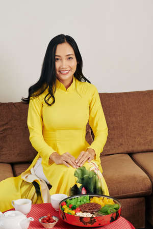 Young Asian woman in traditional dress sitting on sofa in front of table with tea and mut plate served for Tetの写真素材