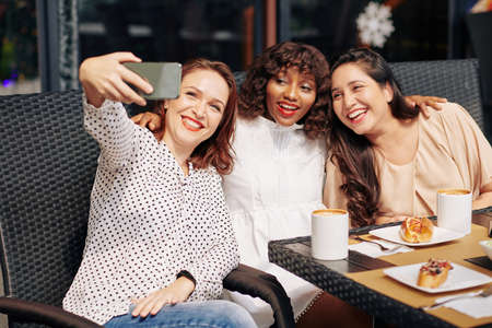 Multi-ethnic happy group of friends sitting at cafe table and posing for selfieの写真素材