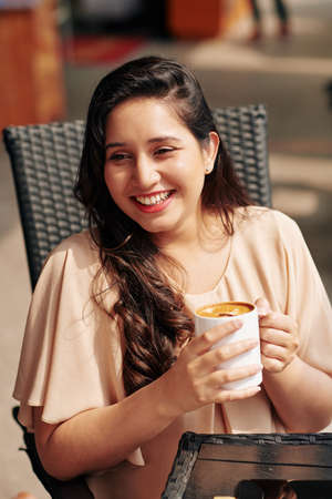 Portrait of young laughing beautiful woman drinking mug of delicious cappuccino in cafeの写真素材