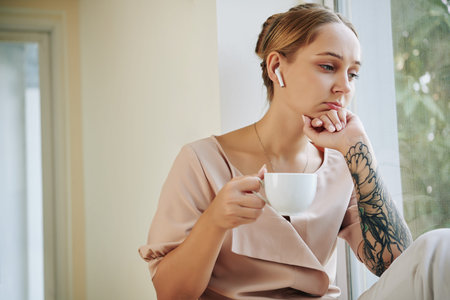 Young woman listening to serious audiobook in her earbuds when sitting on window sill and drinking...