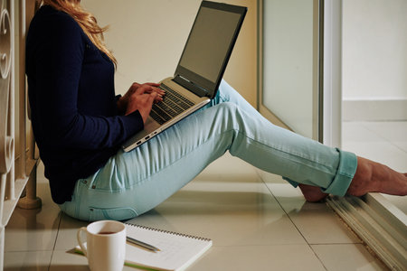 Cropped image of young woman sitting on the floor at home, working on laptop or doing homework for...