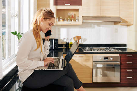 Young positive businesswoman sitting on kitchen counter, reading e-mails on laptop and talking on phone with clientの写真素材