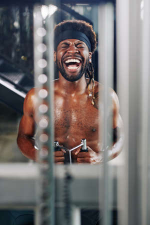 Vertical portrait of African American man laughing at something while doing exercise using fitness machine in gymの写真素材