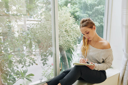 Horizontal shot of romantic Caucasian girl sitting on window sill writing somethjing in her notebook