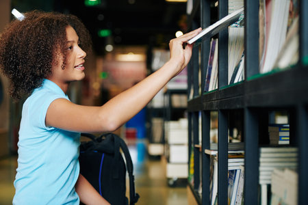 Horizontal side view waist up shot of teenage girl taking book from shelf in modern bookstore