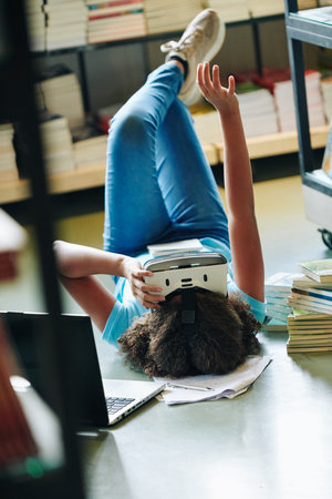 Relaxed teen girl having fun using augmented reality technologies in modern library, vertical shot