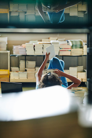 Vertical through bookshelf shot of unrecognizable girl lying on floor using modern virtual reality...