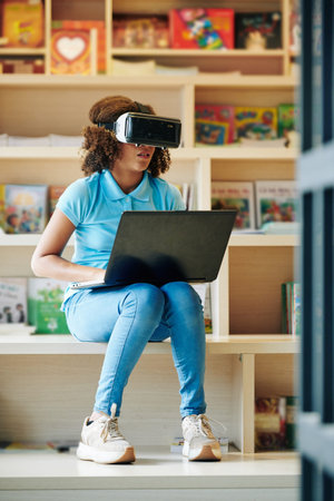 Teen girl wearing casual clothes sitting in modern school library using VR glasses and laptop...