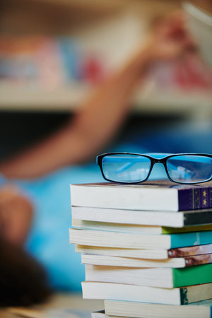 Vertical shot of eyeglasses on stack of books with unrecognizable girl blurred on background
