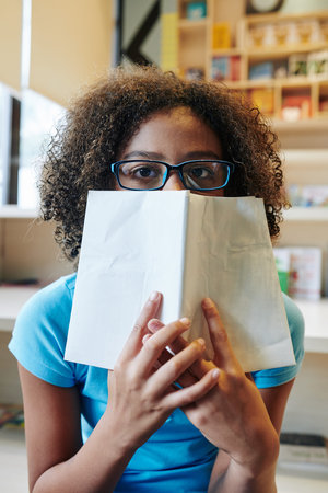 Vertical medium portrait of girl with curly hair wearing eyeglasses covering half of her face with...