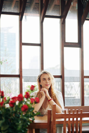 Attractive young woman with wavy blond hair sitting alone in modern restaurant looking away thinking...