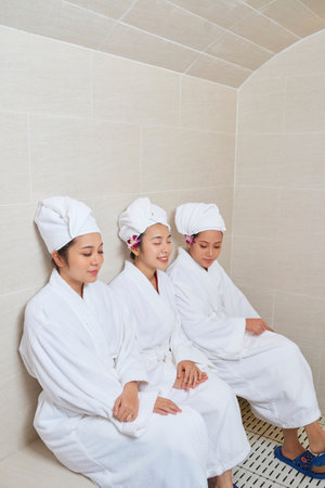 Three Asian women wearing white robes and towel turbans sitting together relaxing in spa salon, vertical shotの写真素材