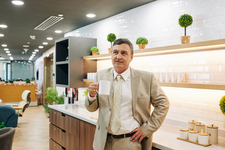 Middle-aged businessman standing in office kitchen holding cup of coffee looking away thinking about...