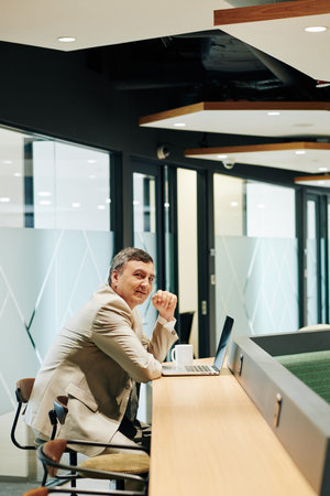 Vertical shot of mature Caucasian businessman working alone in modern office looking at camera
