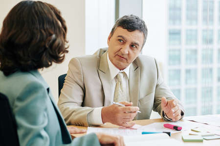 Caucasian businessman wearing beige suit sitting at desk in front of his unrecognizable client working with herの写真素材