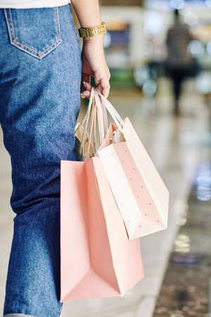 Unrecognizable woman wearing jeans walking along mall with paper shopping bags, vertical shotの写真素材