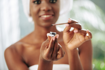 Cheerful young woman holding make up brush and eye shadow looking at camera smiling, horizontal...