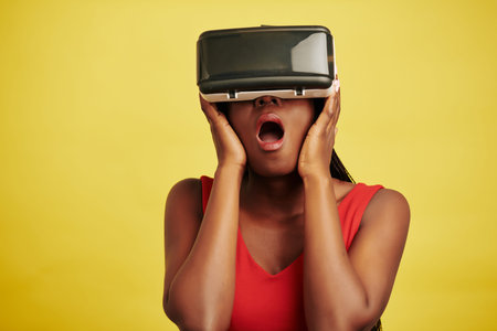 Horizontal studio portrait of Black woman wearing VR headset for first time being impressed, yellow...