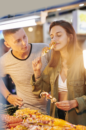 Vertical medium portrait of young Caucasian woman enjoying Asian street food while her boyfriend looking at her and telling her somethingの写真素材
