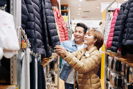 Horizontal shot of young Asian couple choosing casual down jacket for woman in modern clothes shopの写真素材