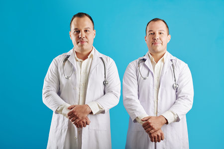 Horizontal studio portrait of twin brothers working as doctors standing against blue wall backgroundの写真素材