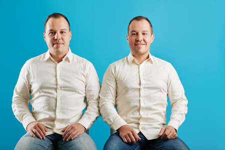 Horizontal studio portrait shot of two twin brothers sitting against blue wall background looking at cameraの写真素材