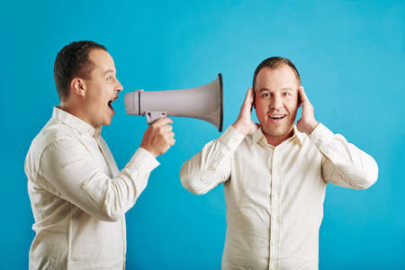 Horizontal medium shot of man with megaphone shouting loud at his twin brother, blue backgroundの写真素材