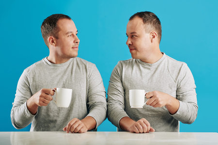 Young adult male twin siblings sitting at table looking at each other holding cups with coffee, blue backgroundの写真素材