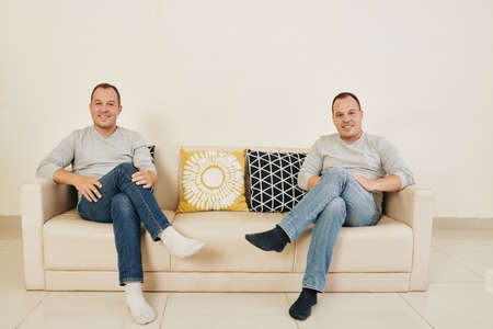 Horizontal shot of young adult twin brothers sitting together on sofa with legs crossed looking at camera smilingの写真素材