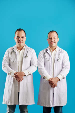 Vertical studio portrait of twin brothers working as doctors standing against blue wall backgroundの写真素材