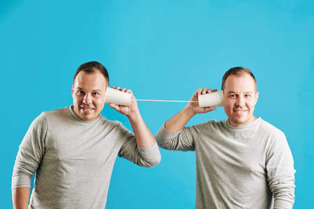 Young adult Caucasian twin brothers playing with plastic cup telephone looking at camera, blue wall backgroundの写真素材