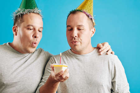 Horizontal studio portrait of young adult twin brothers standing together with small piece of birthday cake blowing out candleの写真素材