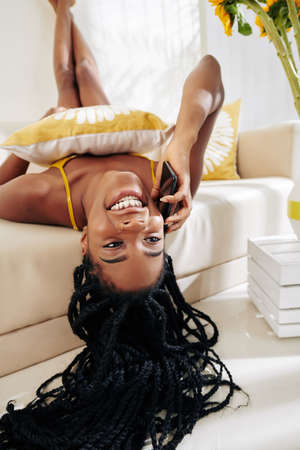 Laughing young Black woman lying on sofa with head hanging upside down and calling on phoneの写真素材
