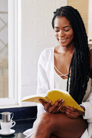 Smiling charming young Black woman drinking morning coffee and reading a book