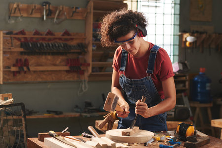 Concentrated curly woman in ear defenders and goggles using hammer and chisel while working with...