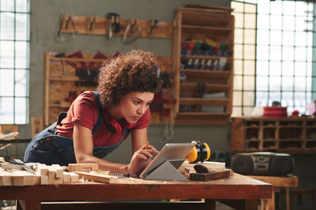 Young concentrated woman with curly hair reading instructions on digital tablet before working with woodの写真素材