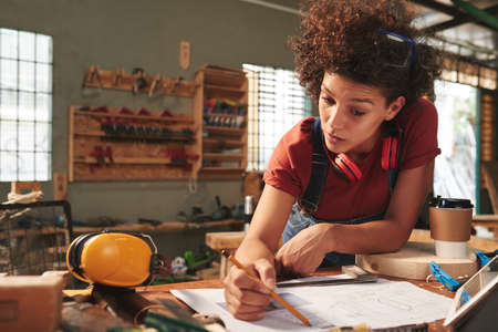 Young attractive female carpenter thoroughly checking measurements on blueprint before working with woodの写真素材