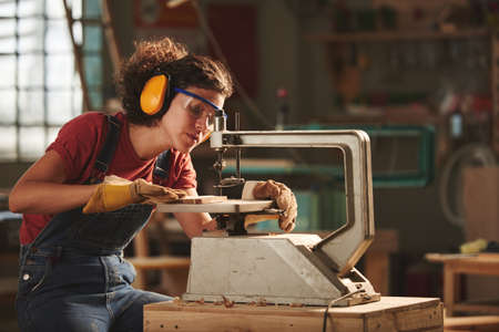 Young concentrated woman in protective eyewear and earmuffs making holes in wooden plank with drill pressの写真素材