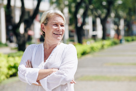 Positive mature woman in silk white blouse folding arms when standing outdoors on sunny day