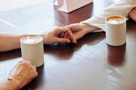Close-up image of young woman touching hand of her mother to support her when drinking coffee in...