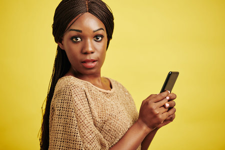 Horizontal studio portrait of attractive Black woman holding smartphone looking at camera, yellow...