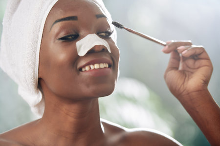 Horizontal head and shoulders portrait of young African American woman applying mascara on her...