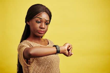 Horizontal studio portrait of Black woman getting late for something checking time, yellow backgroundの写真素材